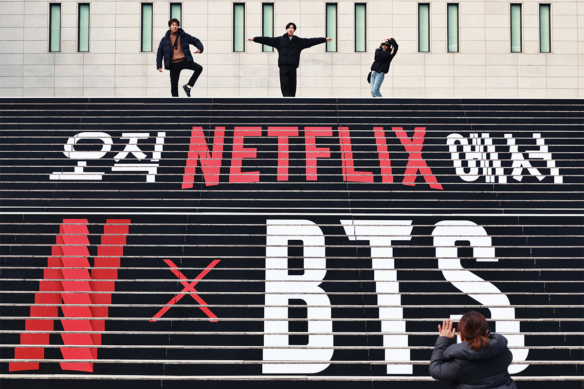 سياح أجانب يلتقطن صورة أمام شعار حملة ترويجية لعودة فرقة بي تي إس على سلالم مركز سيجونغ للفنون الأدائية في حي جونغنو-غو، سيئول في يوم 11 مارس حيث من المقرر أن تعود بي تي إس بكامل أعضائها بألبوم جديد في يوم 20 مارس.