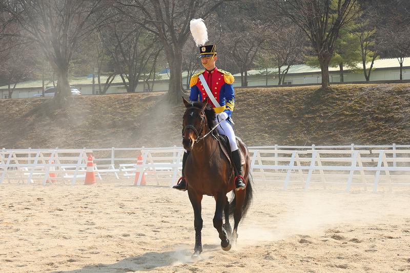 الطالب الضابط هو جون-سوك (السنة الرابعة) في غمرة تدريباته في ميدان الفروسية. هنا، لا ترسم المسارات الدقيقة والتوازن المذهل إلا نتيجة دأبٍ مستمر وتواصلٍ وجداني عميق؛ فالحصان لا يرضخ للأمر المجرد، والفارس لا يقود بالقوة فحسب، بل يكتمل التدفق كمسار واحد من خلال قراءة كل منهما لإيقاع الآخر وتوافق أنفاسهما معًا. وعند اللحظة التي يتحرك فيها الإنسان والحيوان ككيان واحد، يتجلى الانضباط والتحكم بوضوح أكثر.