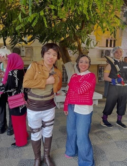 الصورة تظهر لقطة عفوية لرحاب رمضان ببلوزة حمراء مشغولة وهي تبتسم بجانب شاب يرتدي زي الكوسبلاي لشخصية إيرين ييغر الشهيرة من أنمي هجوم العمالقة، في أجواء تجمع بين عشاق الثقافات الآسيوية بمصر. (مصدر: رحاب رمضان)