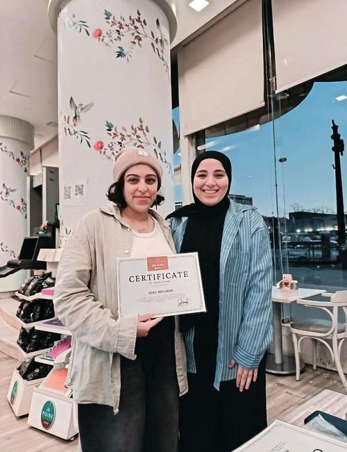 شروق أيمن تقف بجانب إحدى الطالبات التي تحمل بفخر شهادة إتمام الدورة التدريبية باللغة الكورية داخل مقهى هادئ. (المصدر: شروق أيمن)