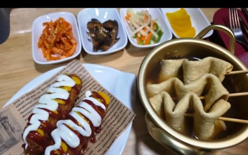 مجموعة من الأطباق الكورية تضم «الكورن دوغ» المغطى بالصلصة، وأعواد «الأودينغ» (كعك السمك الكوري)، إلى جانب مجموعة متنوعة من المقبلات. (الصورة من فاطمة مسعد، بعد الحصول على إذن بالنشر)
